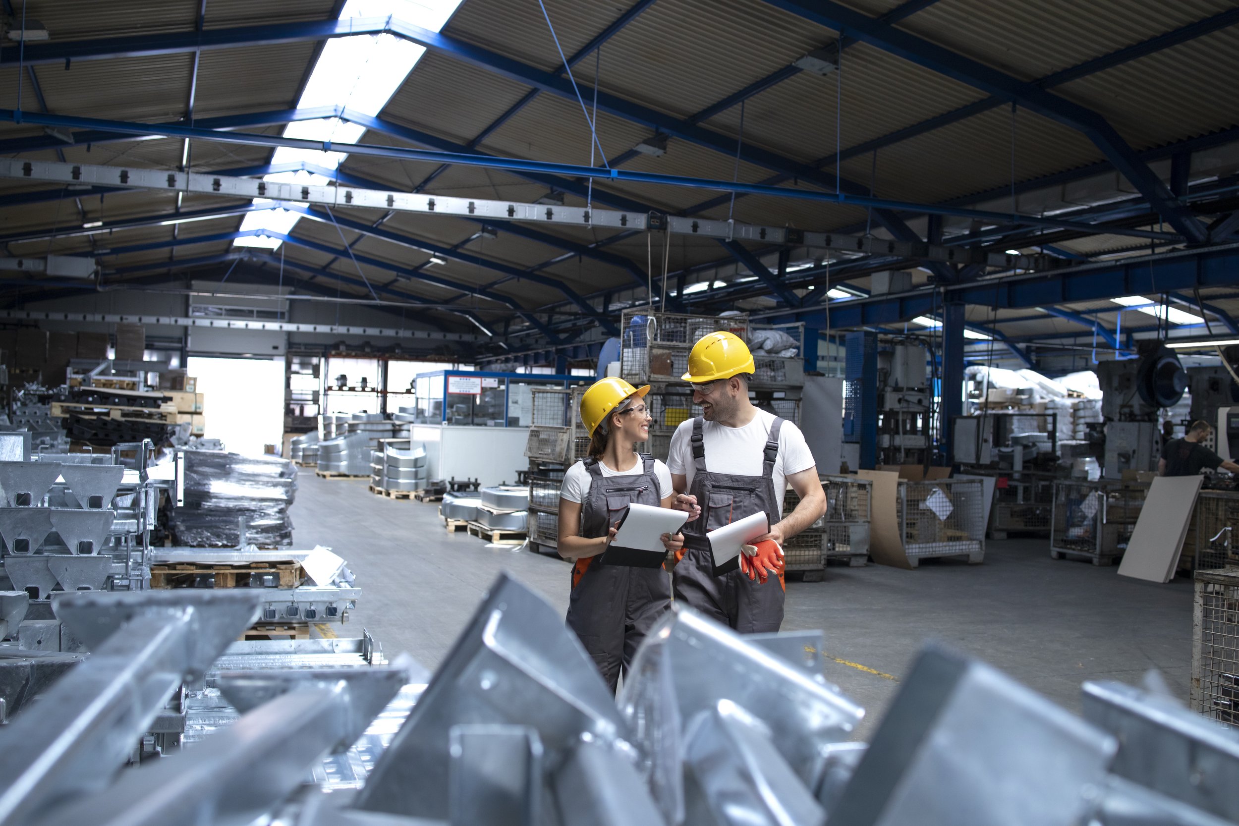 Twee vakbekwame werknemers in een grote fabriek met helmen die praten, terwijl ze documenten en tablets vasthouden, omgeven door industriële metalen onderdelen en machines.
