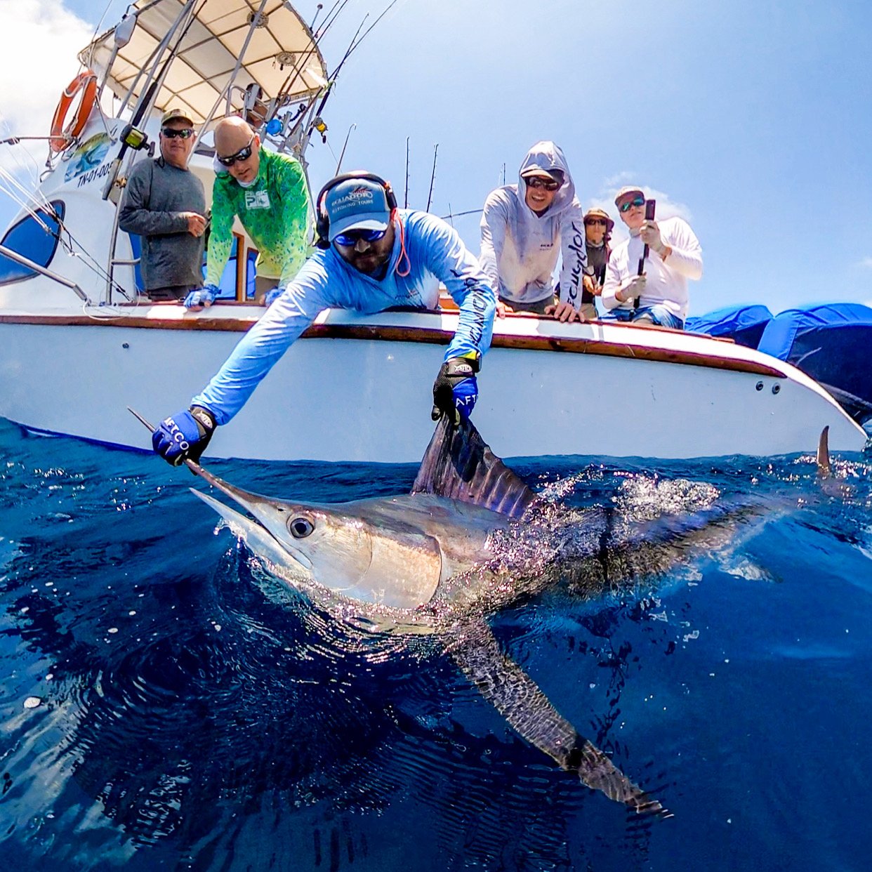 Group of people on a boat catching a large marlin fish in the ocean on a sunny day.