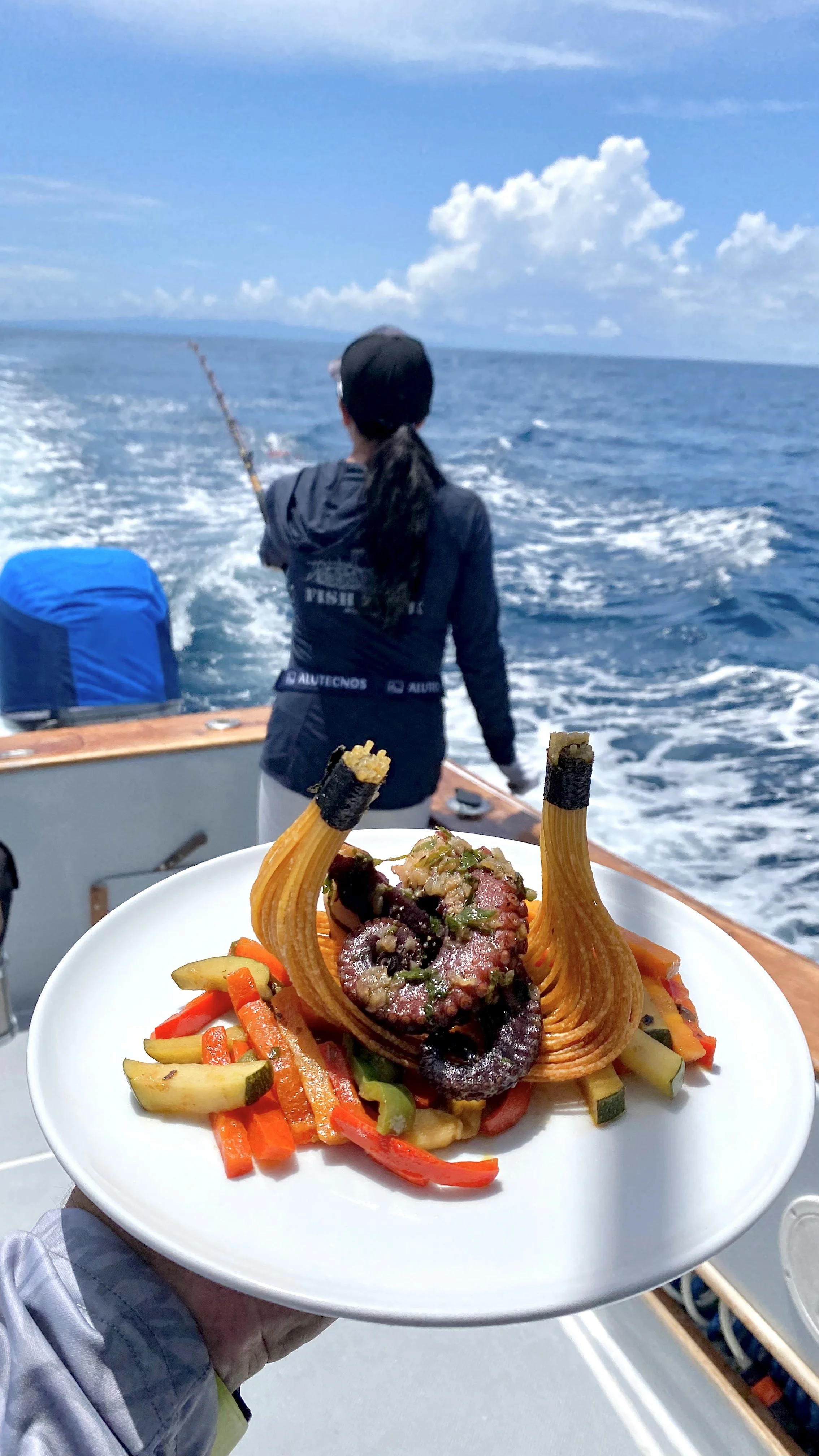 A plate with a gourmet dish of spaghetti with a seared tuna steak, garnished with vegetables, held up on a boat in the ocean with a person fishing in the background.