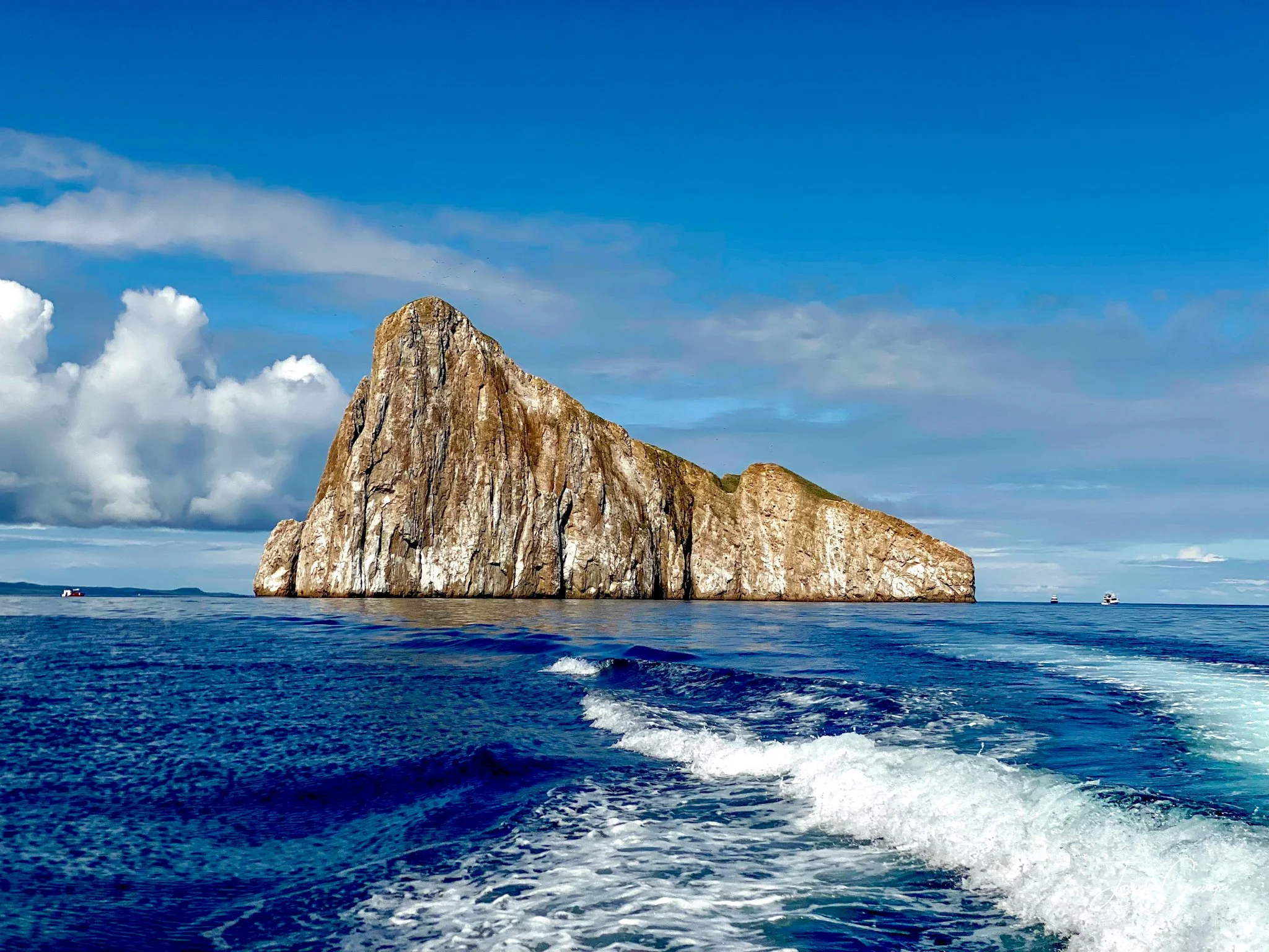 A large rocky island formation rises from the ocean, with a boat trail in the water and a partly cloudy blue sky above.