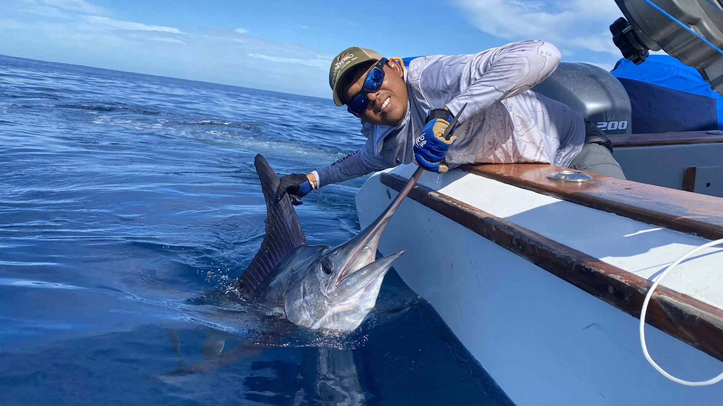 Man in sunglasses and cap holding a large fish over the side of a boat in the open ocean.