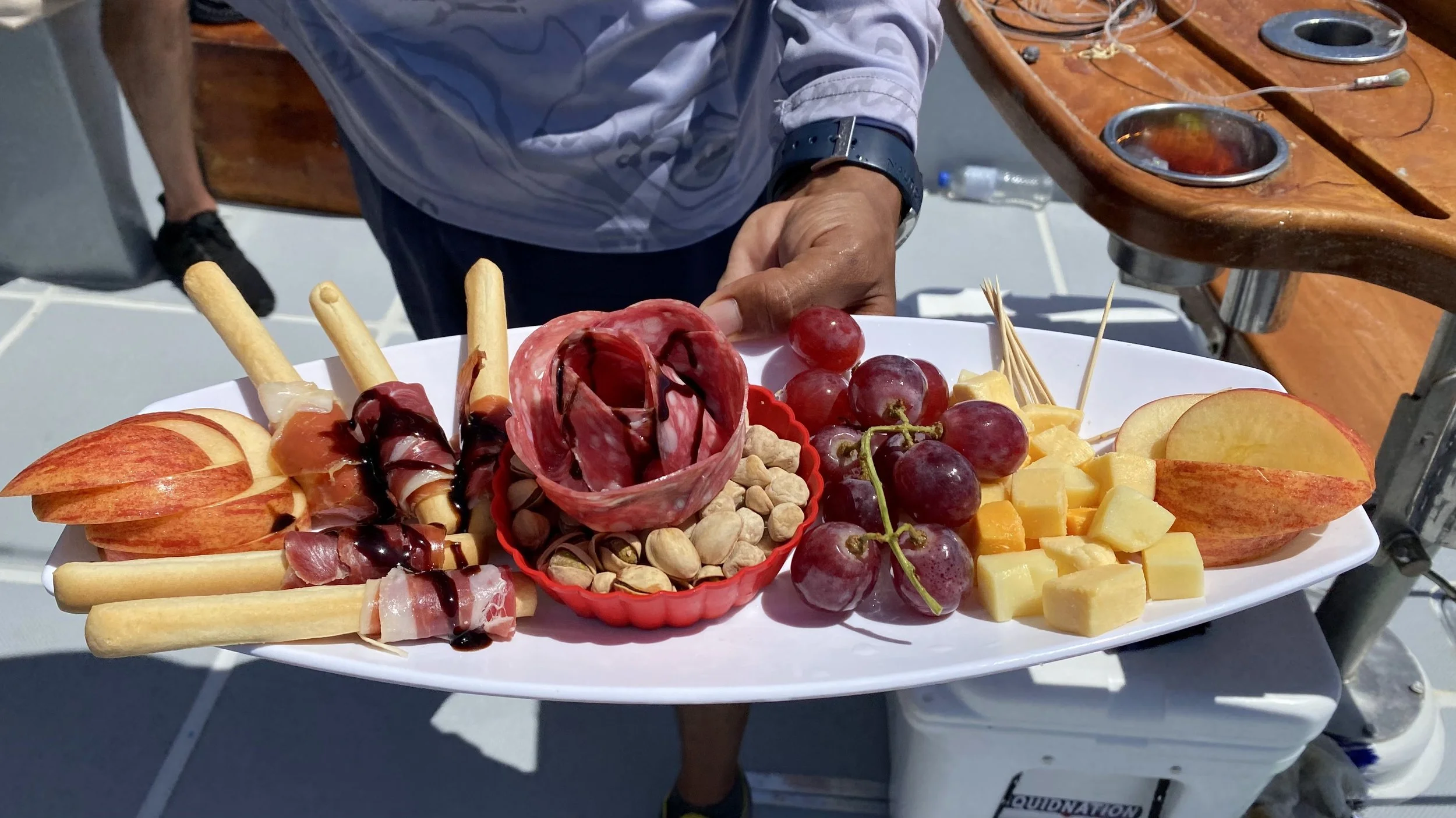 A person holding a white platter with apple slices, grapes, cheese cubes, pistachios, rolled cured meats, breadsticks, and a small bowl of pistachios and chocolate drizzle.
