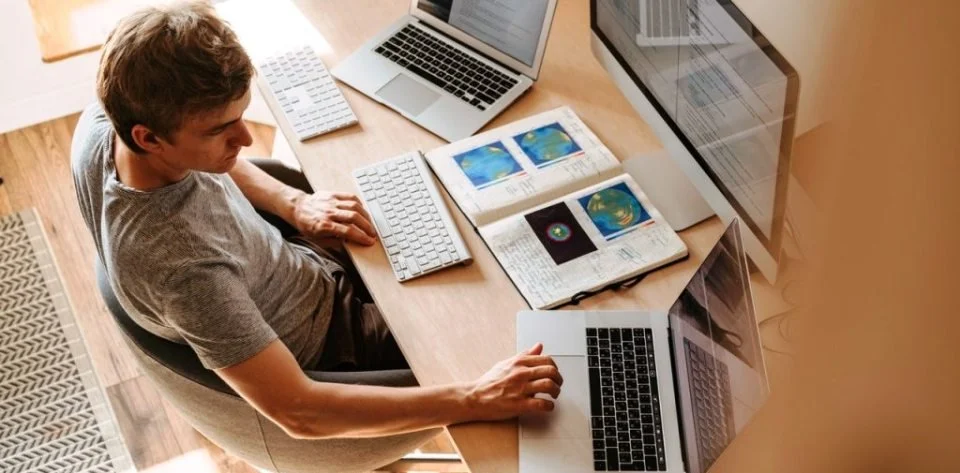 A man working at a desk with multiple computers, a keyboard, journals, and maps open on the desk.