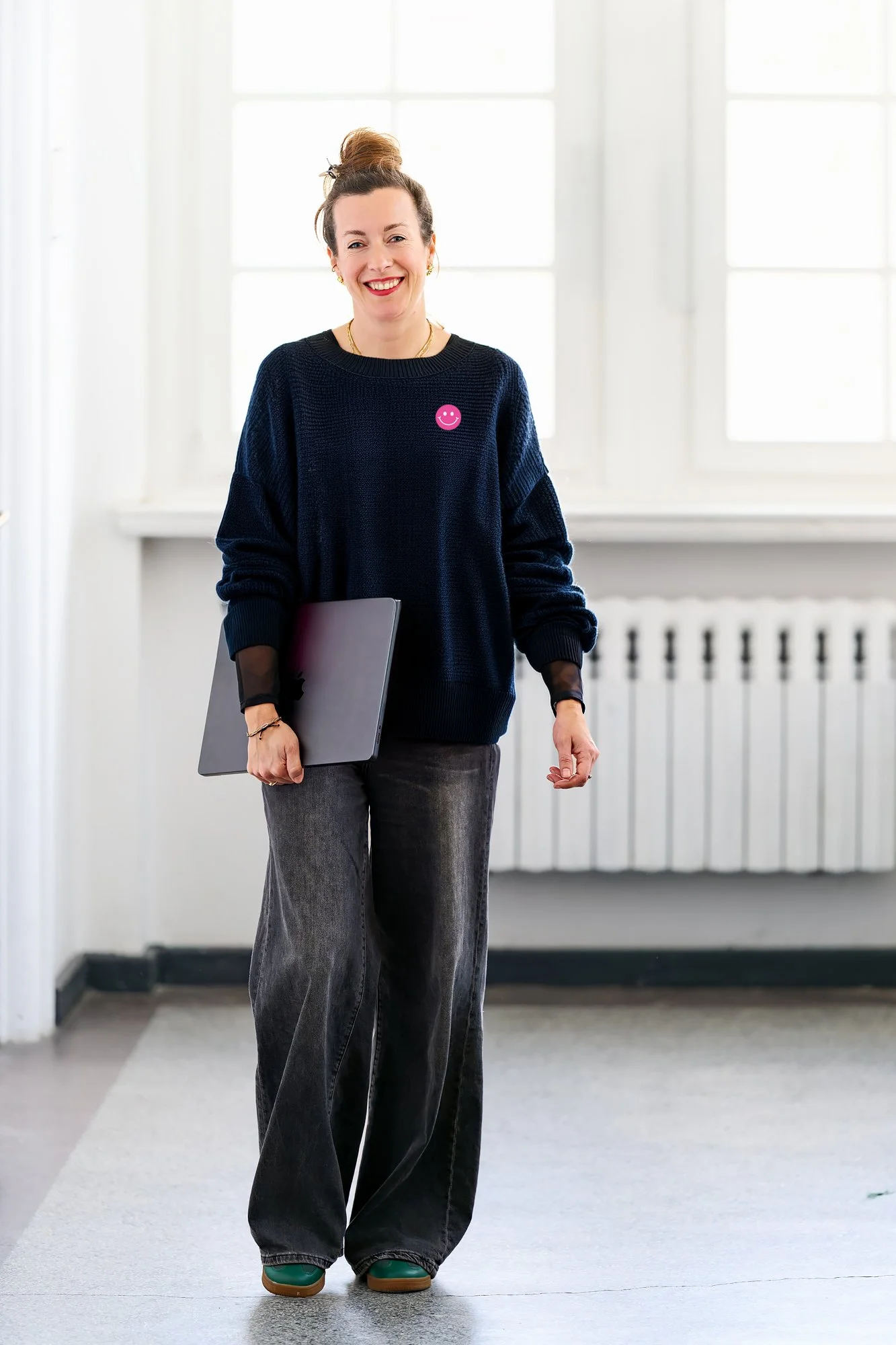 Maria Weding steht in einem hellen Raum, trägt eine dunkle Strickjacke mit einem pinken Smile-Gesicht Button, dunkelblauem Pullover, hält einen Laptop und lächelt.