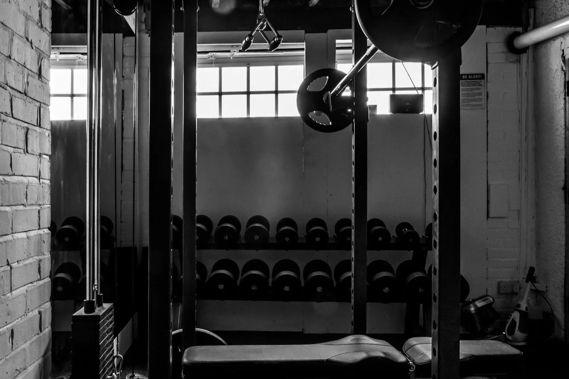 A monochrome image of a gym with a squat rack, dumbbells on racks, and fitness equipment near a brick wall and a window.