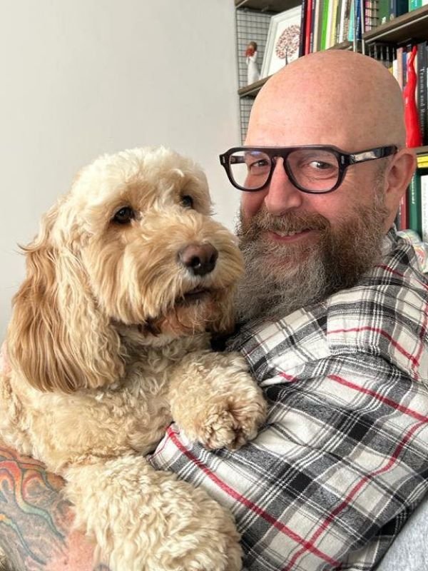 Photo of Ben Perry sitting in an armchair holding his Labradoodle, Elliot