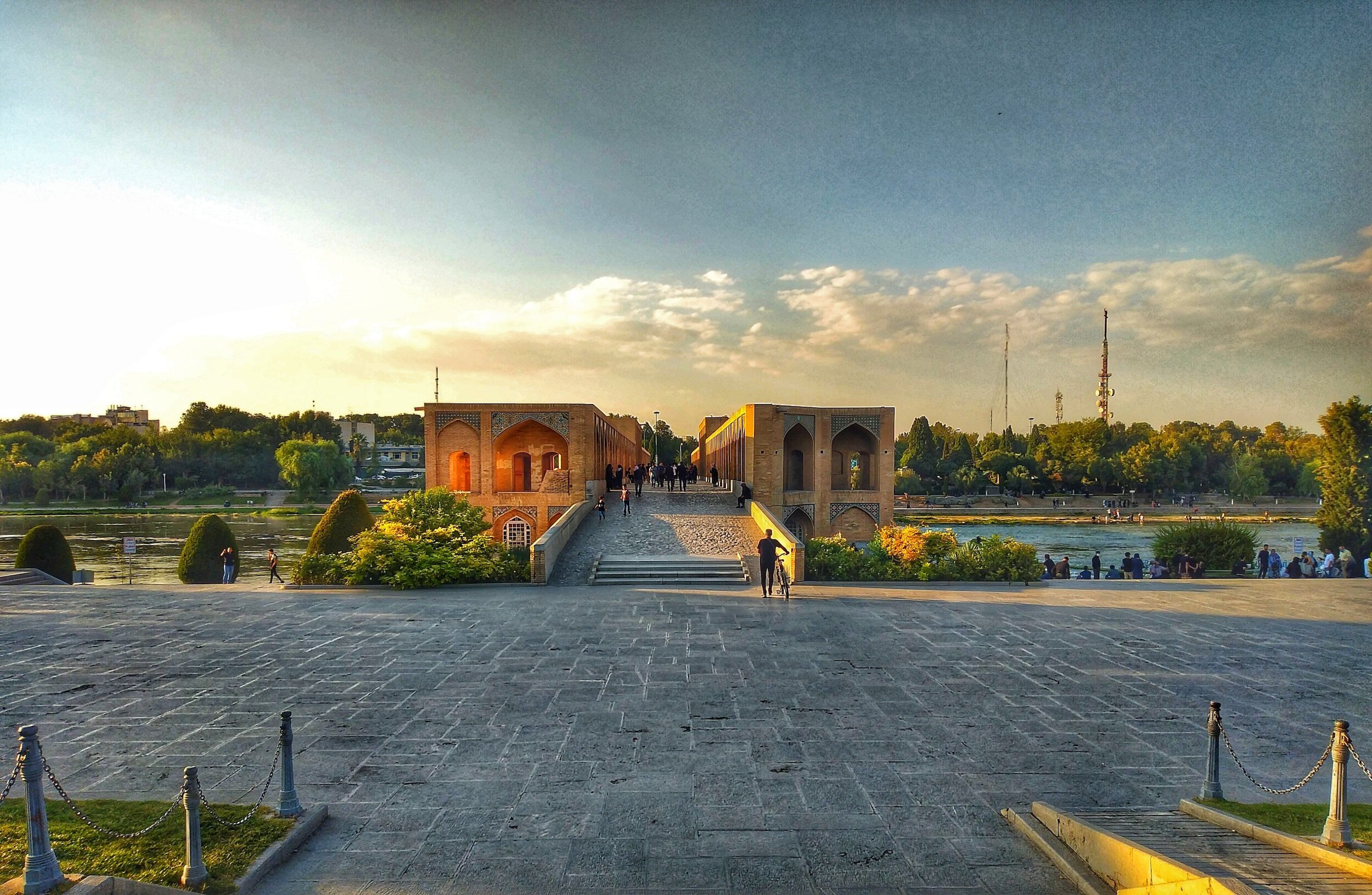 Sunset view of historic bridge over a river with trees and city skyline in the background.