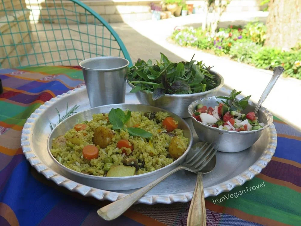 Vegan meal on a metal tray with rice and vegetables, a salad, a bowl of fresh greens, a metal cup, and cutlery, outdoors with plants and flowers in the background.