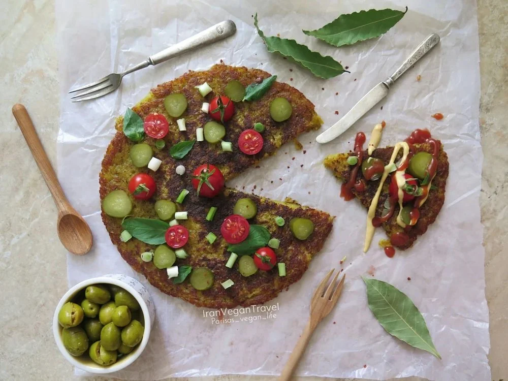 A vegan breaded cutlet with cherry tomatoes, green olives, chopped green onions, and fresh basil leaves, garnished with sauces and served on parchment paper.