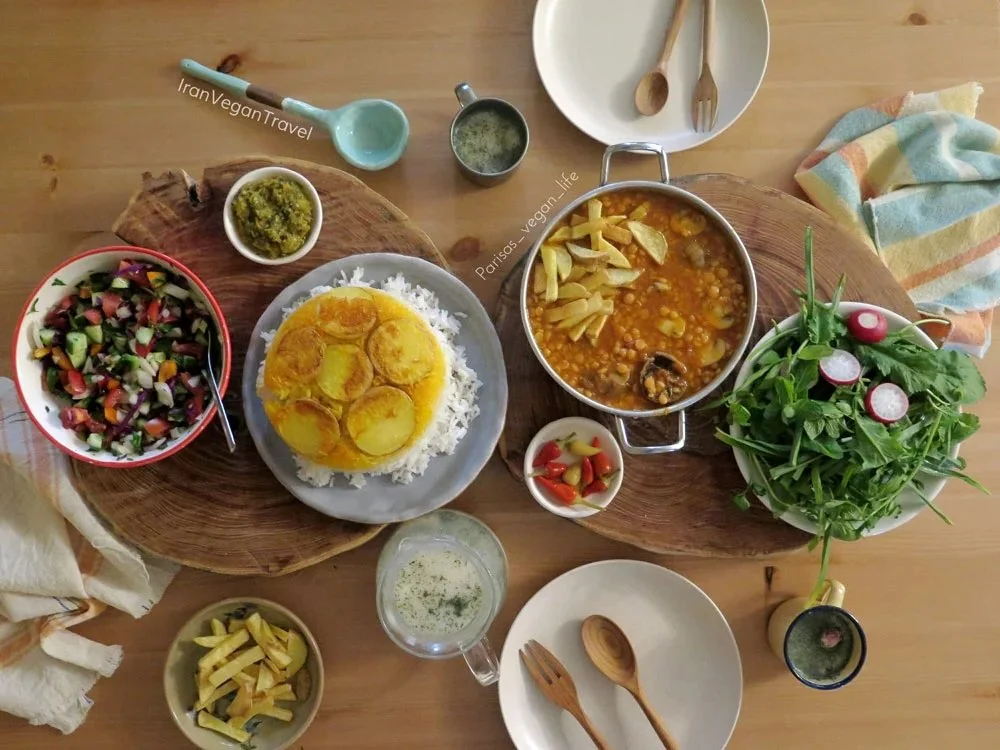 Top-down view of a wooden table with various dishes including a bowl of salad, a plate of rice with yellow curry, a pot of lentil stew, a bowl of pickled vegetables, a small bowl of green sauce, a glass of water, and a bowl of fresh greens with radishes. There are also utensils, a striped towel, and small bowls of pickled vegetables and green sauce.