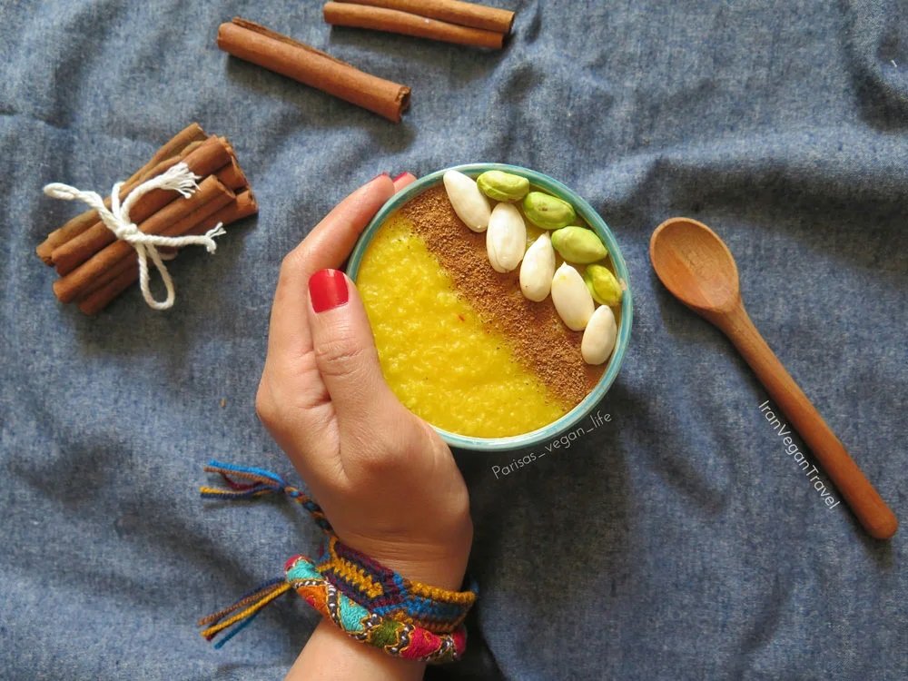A hand holding a bowl of yellow vegan dessert topped with a row of almonds and pistachios. Nearby, there are cinnamon sticks, a wooden spoon, and a blue fabric surface.