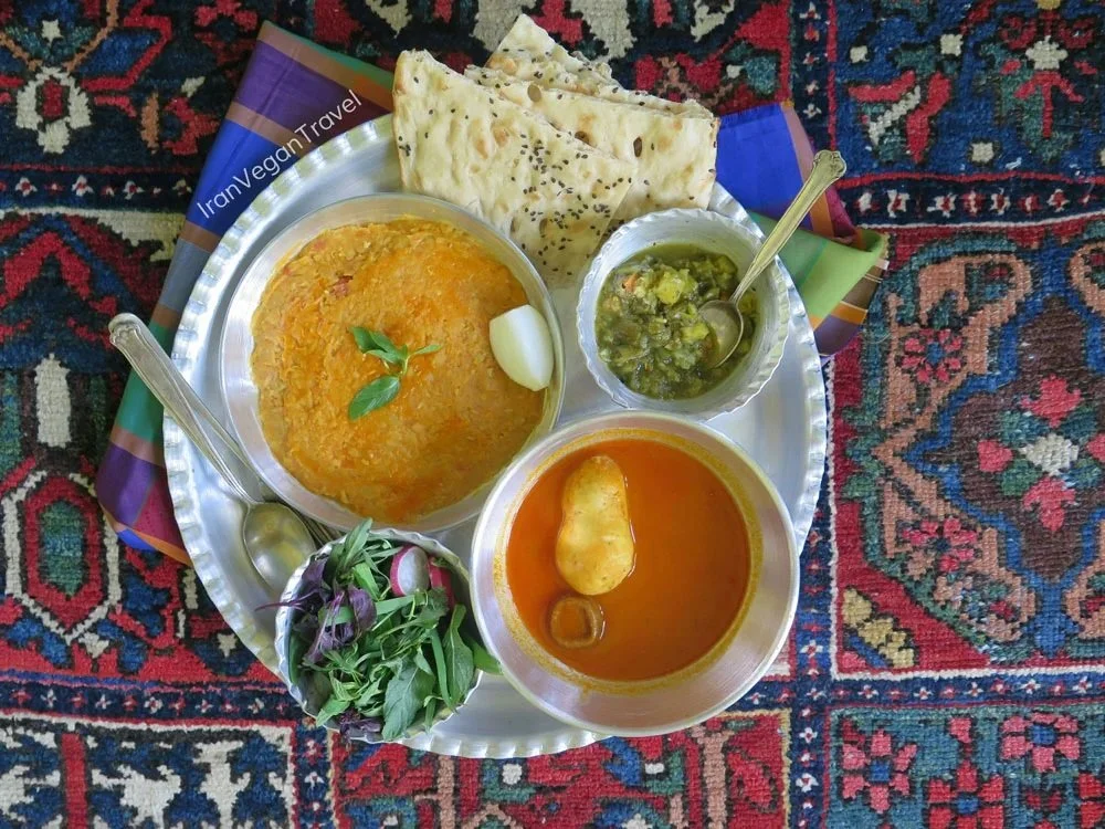 A traditional Iranian vegetarian meal served on a silver tray placed on a Persian rug. It includes a bowl of orange lentil stew garnished with basil and a boiled onion, a bowl of green vegetable relish with spoon, a bowl of tomato-based soup with a potato, a small salad of various greens, and flatbread.