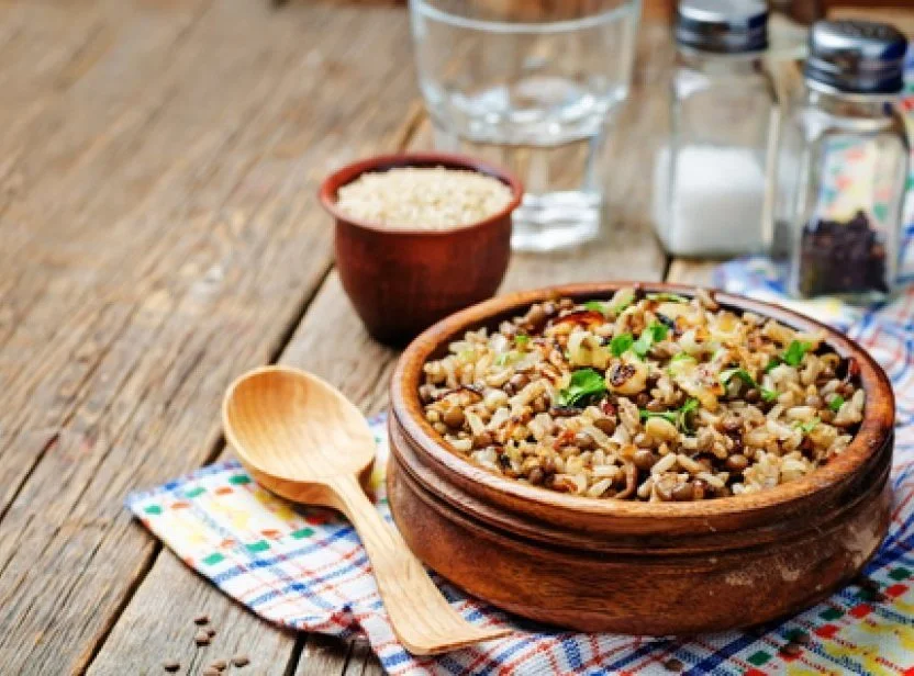 Wooden table with a bowl of rice and a larger bowl of cooked mixed beans and grains, a glass of water, salt and pepper shakers, and a wooden spoon.