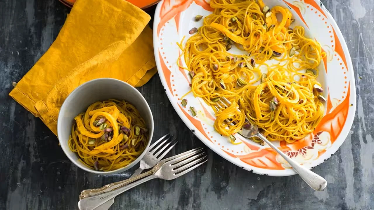 Plate of pasta with yellow noodles and pumpkin seeds, with a smaller bowl of the same pasta, forks, and a yellow napkin on a dark surface.