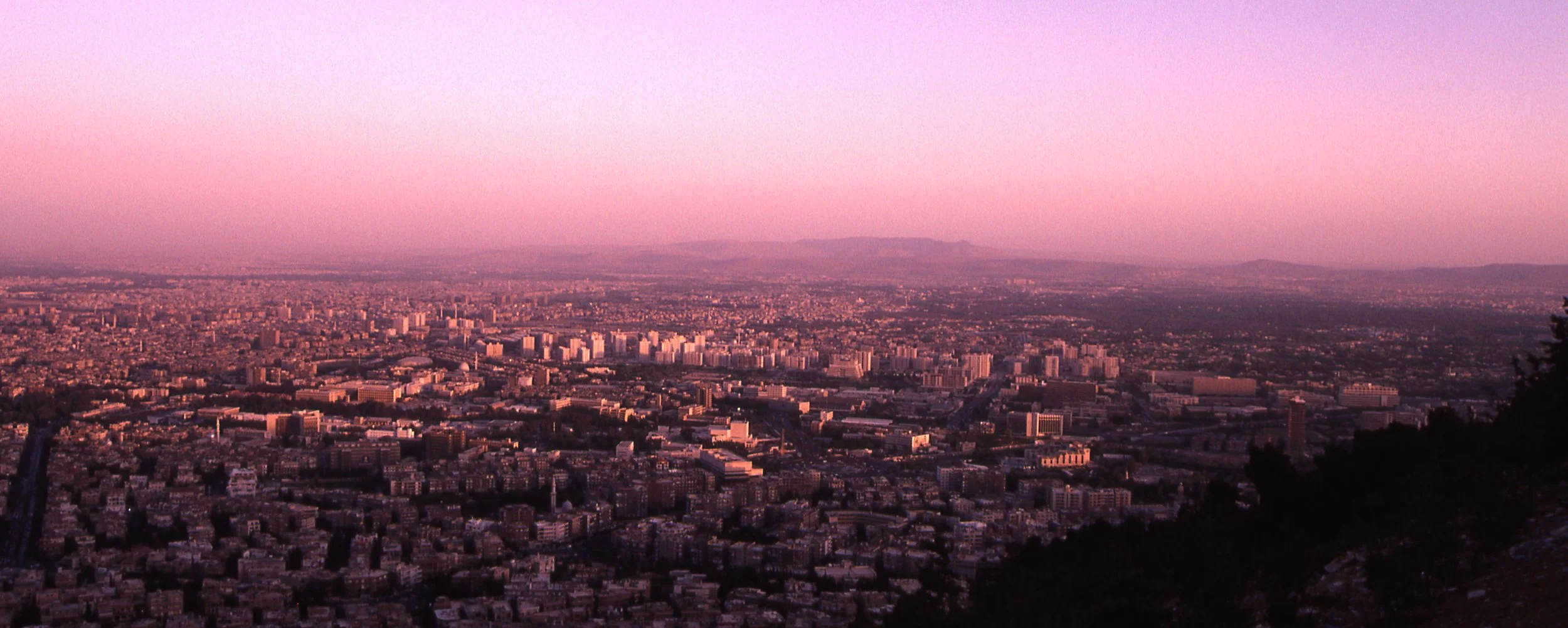 Cityscape at dusk with pink and purple sky over a large urban area with numerous buildings and distant mountains.