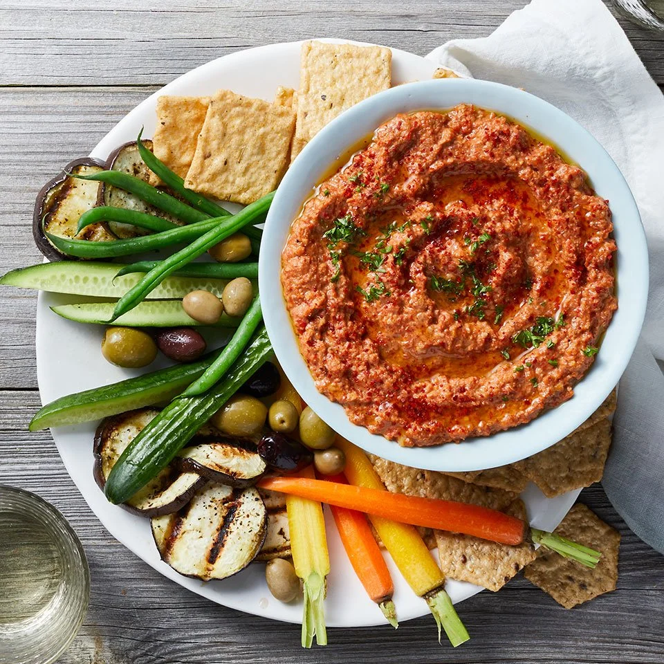 A plate of Mediterranean appetizer with hummus, fresh vegetables, pita chips, and grilled eggplant on a wooden surface.
