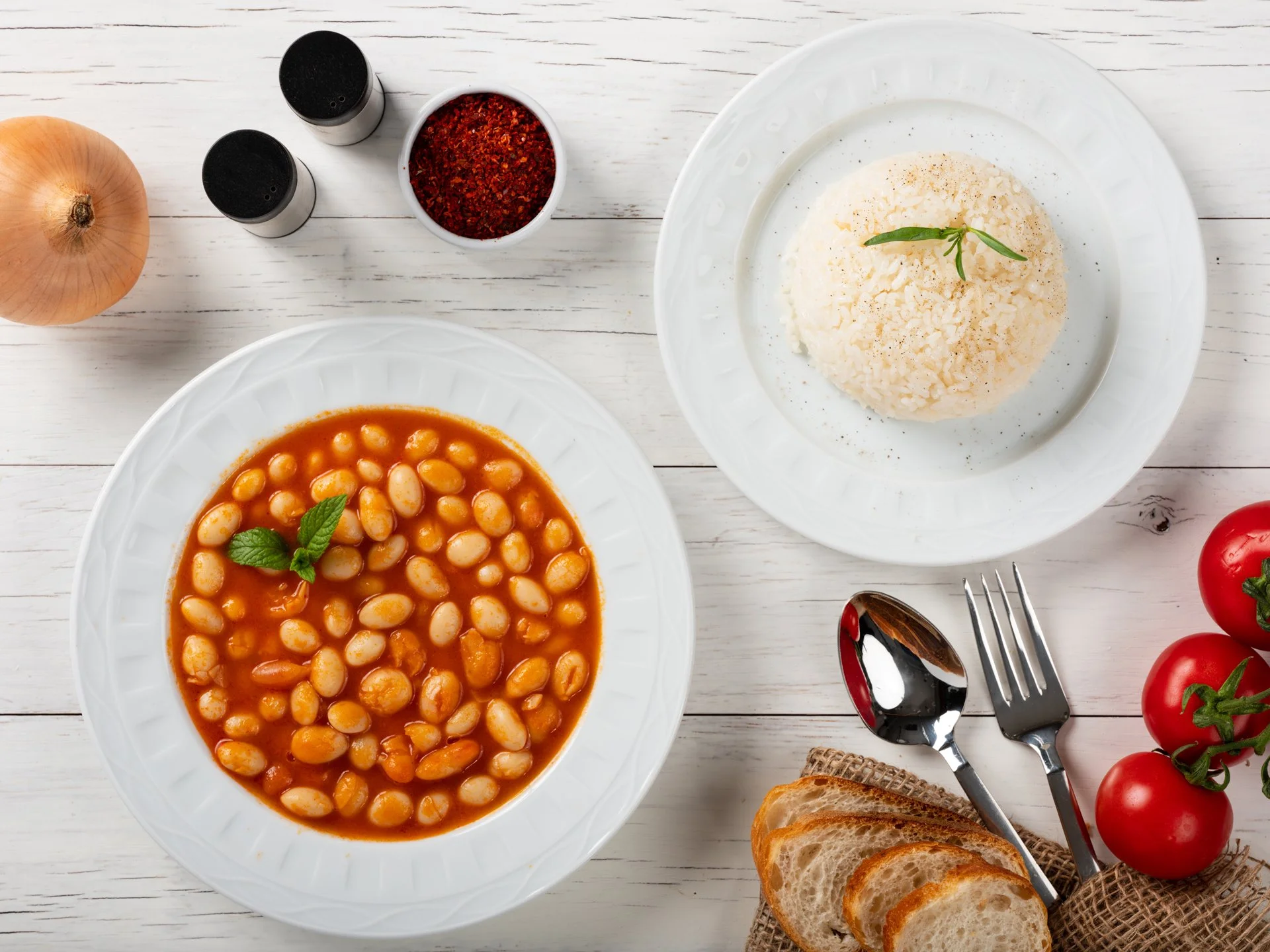Baked beans with a mint garnish on a white plate, white rice with a green herb on a white plate, slices of bread, cherry tomatoes, onion, black pepper, red pepper flakes, spoon and fork on a white wooden table.