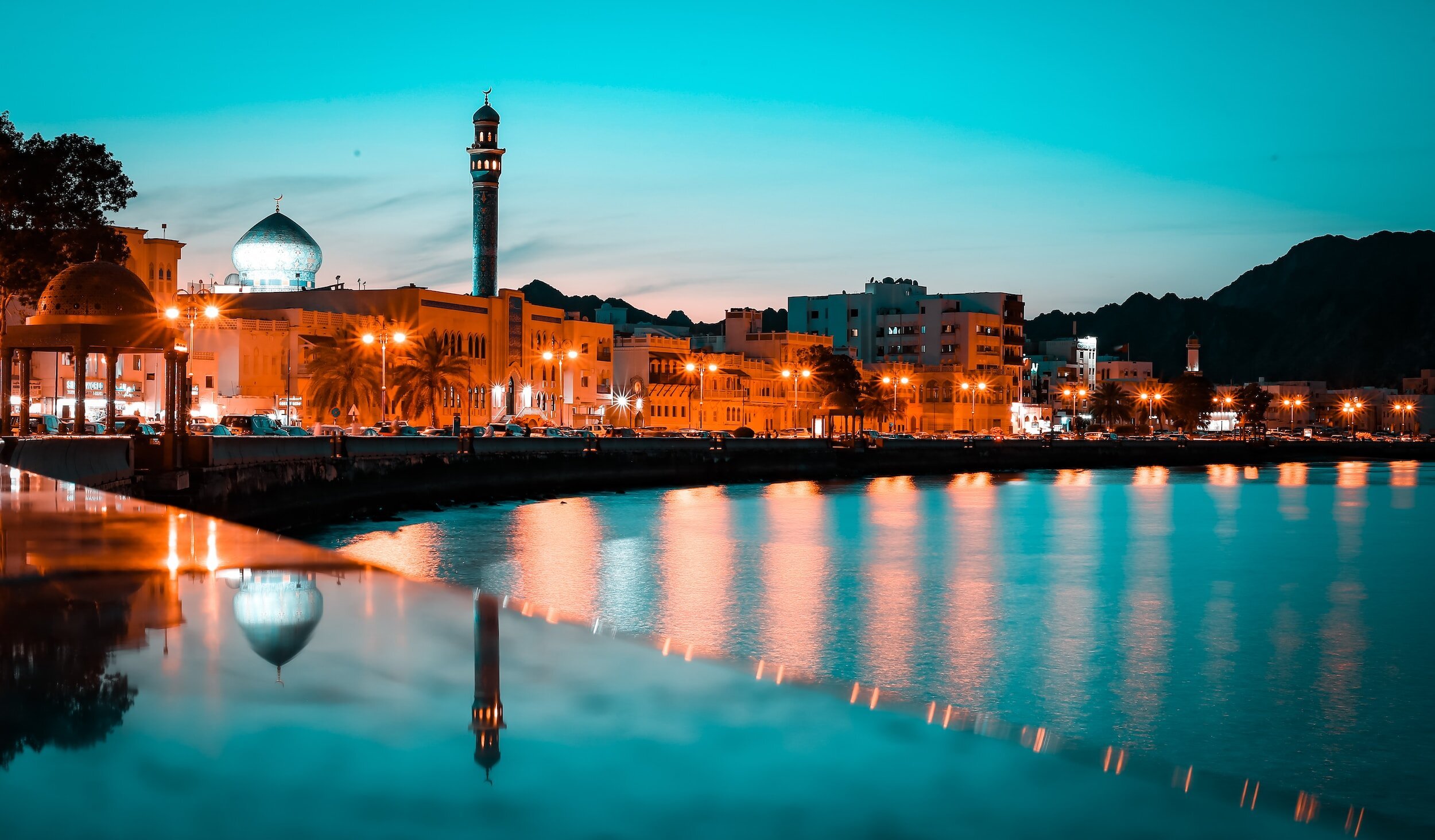 A cityscape at dusk with mosque domes and minaret, waterfront, and illuminated buildings reflected in calm water.