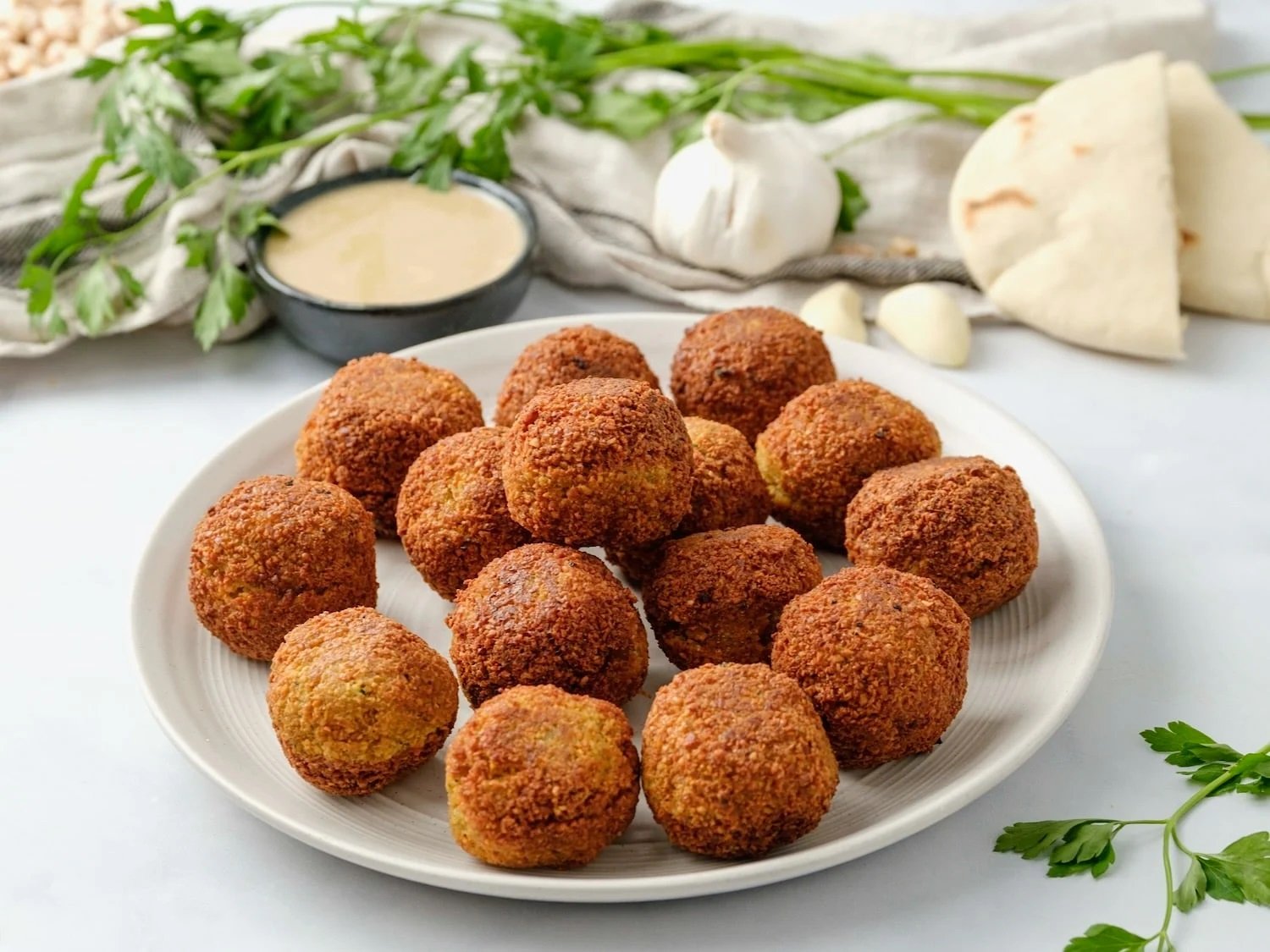 Plate of fried croquettes with a bowl of dipping sauce, garlic, naan bread, and fresh herbs in the background.