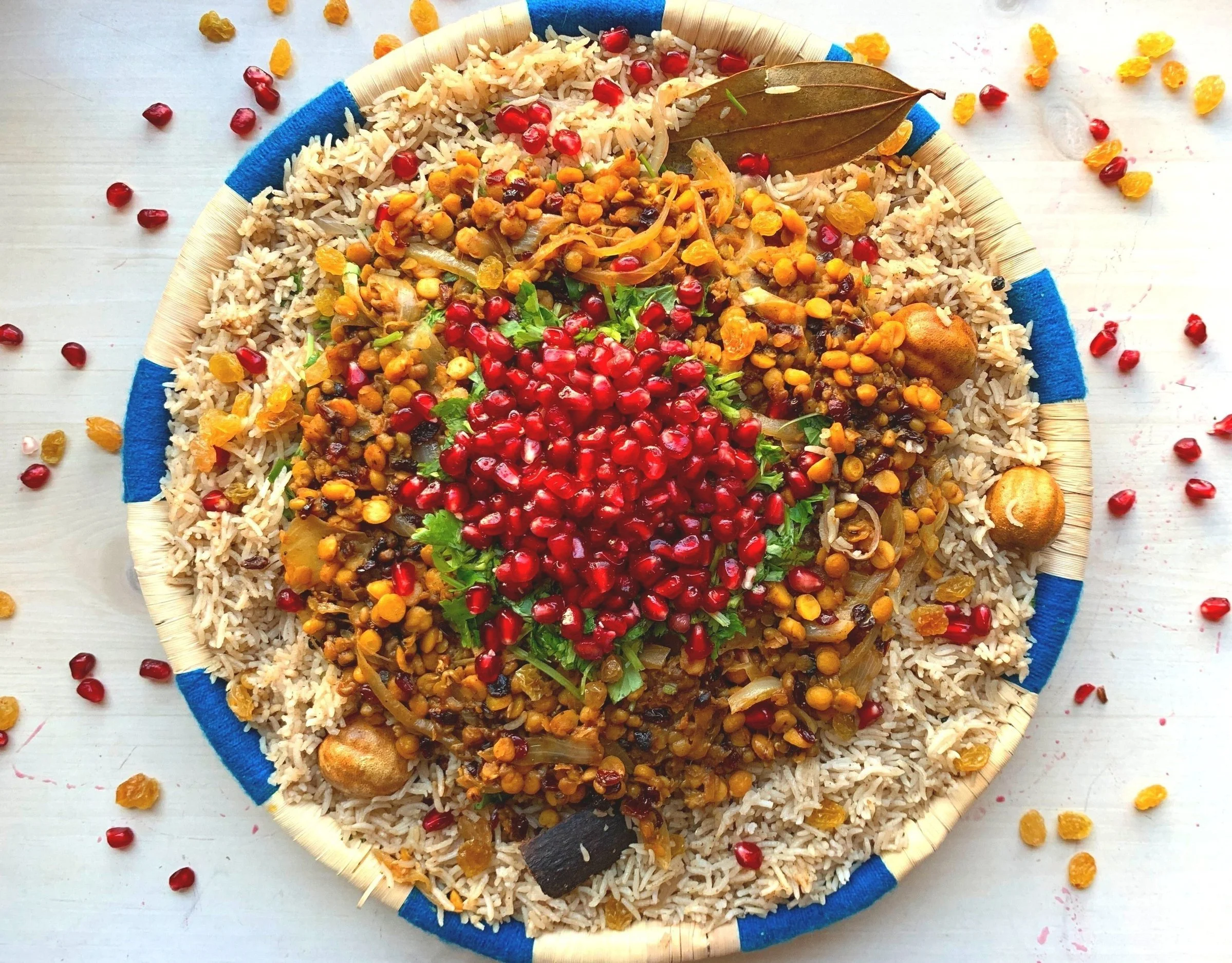 A round woven plate filled with rice, lentils, onions, herbs, pomegranate seeds, and bay leaf, garnished with other spices, and scattered pomegranate seeds and dried raisins around on a white surface.