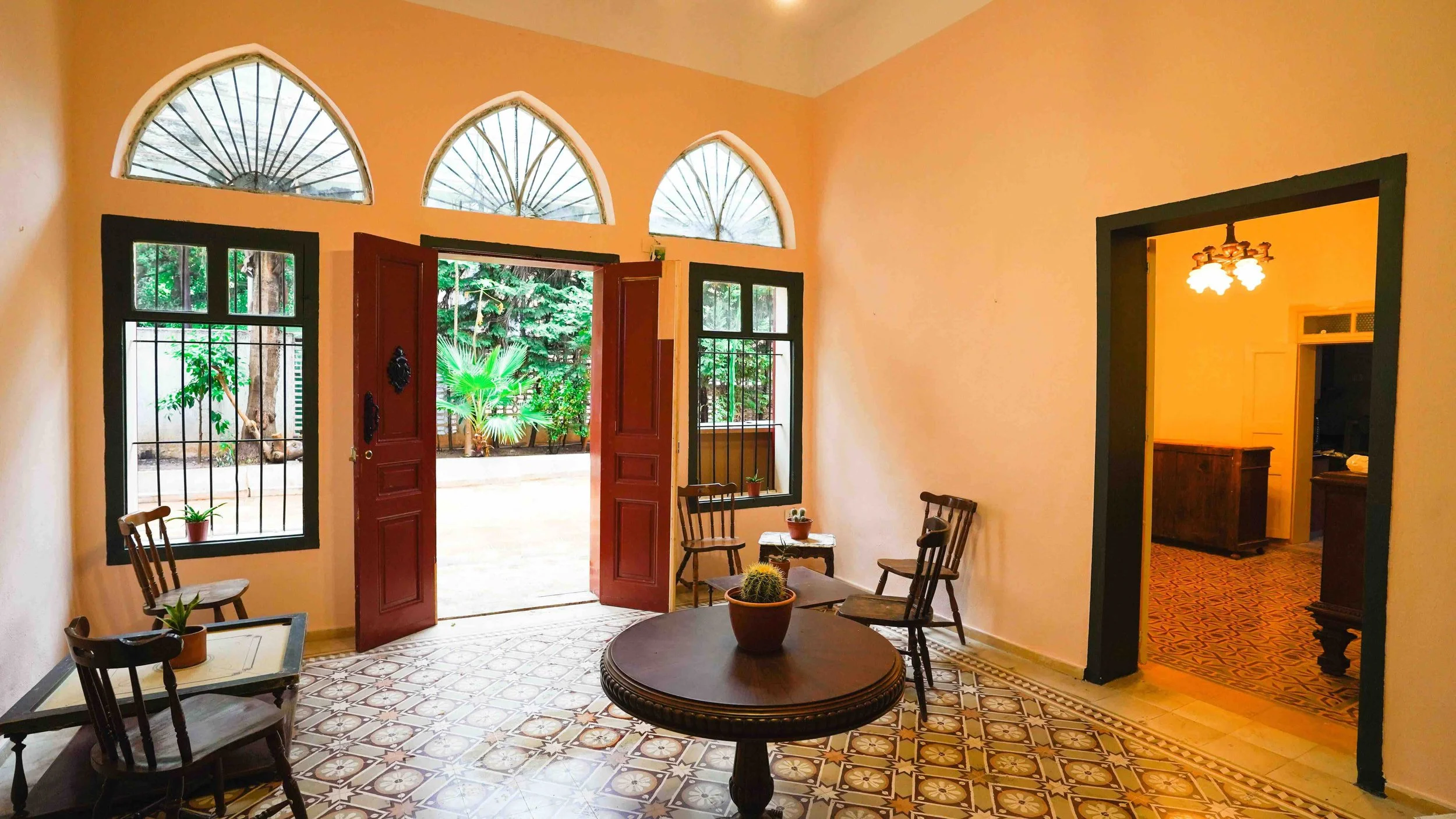 Living room with arched windows, open door to garden, and patterned tile floor, furnished with wooden chairs and potted plants.