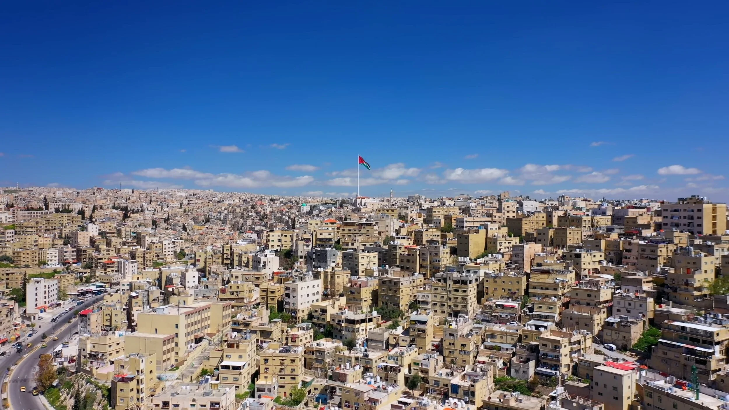 Cityscape of Amman, Jordan with a large Jordanian flag flying on a pole on a hill, under a clear blue sky with some clouds, showing numerous beige and white buildings.
