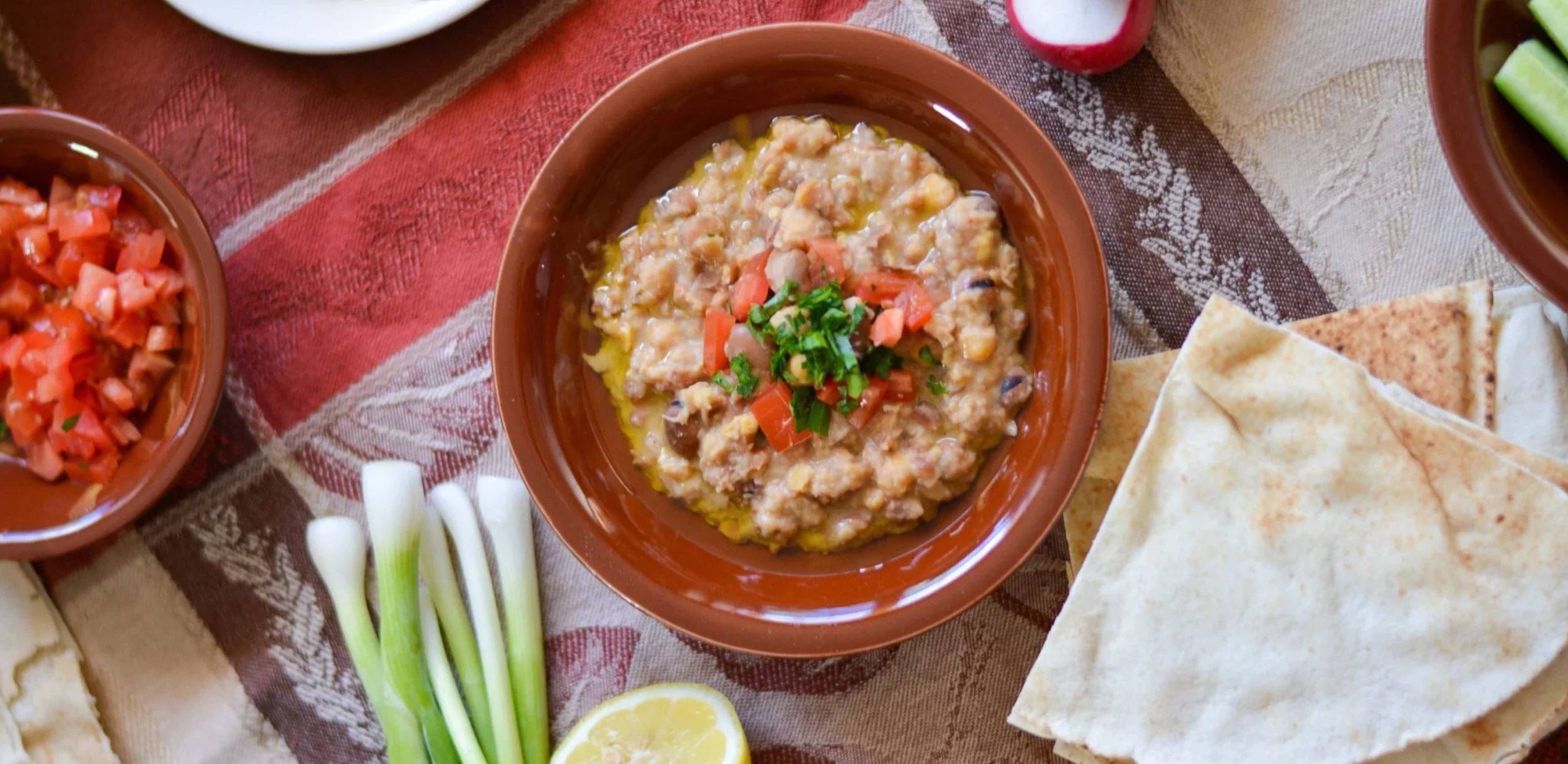 A bowl of Mexican bean dip garnished with chopped tomatoes and cilantro, surrounded by green onions, lemon, chopped tomatoes, and flatbread on a patterned tablecloth.