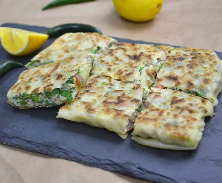 Six slices of pan-fried stuffed flatbread with green peppers, cheese, and meat on a black slate serving board, garnished with lemon wedges and fresh chili peppers, with a lemon in the background.