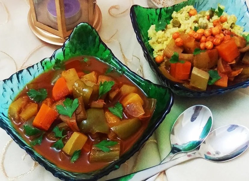 Two glass bowls containing vegetables and grains. The larger bowl has a vegetable stew with carrots, celery, onions, and herbs. The smaller bowl contains mashed potatoes topped with peas, carrots, and gravy. There are two spoons and a candle nearby.