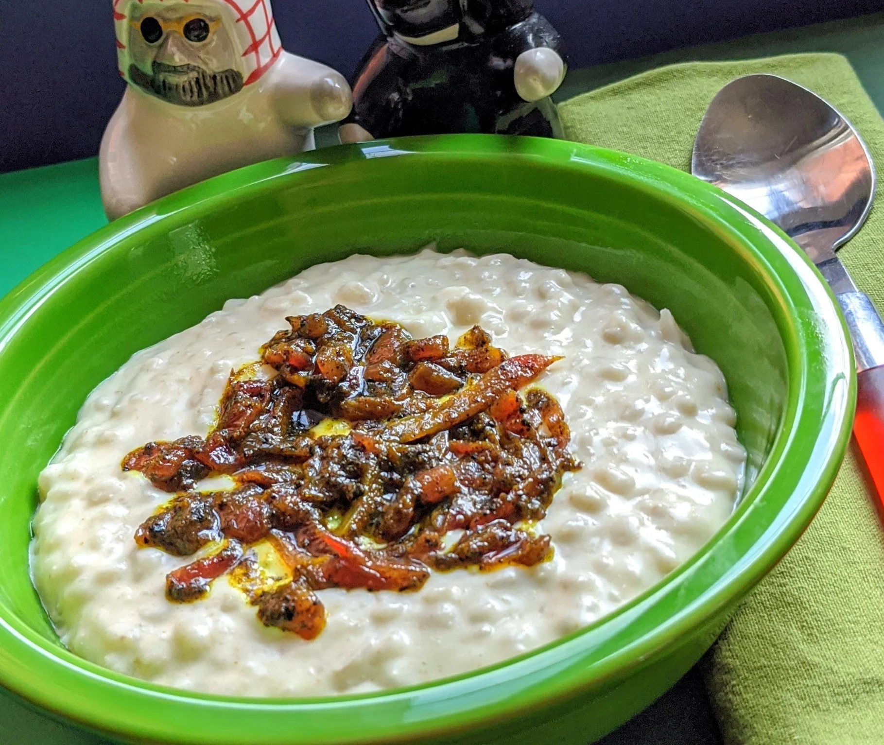 A green bowl filled with creamy rice pudding topped with caramelized fruit or jam, on a green placemat with a large metal spoon nearby, in a setting with ceramic hot dog-shaped salt and pepper shakers in the background.