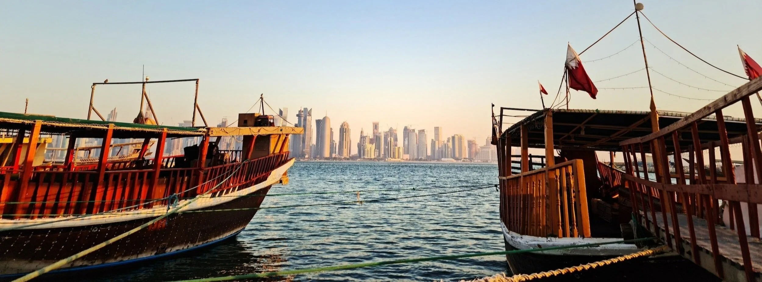 Two wooden boats docked on water with a city skyline in the background, under clear sky.