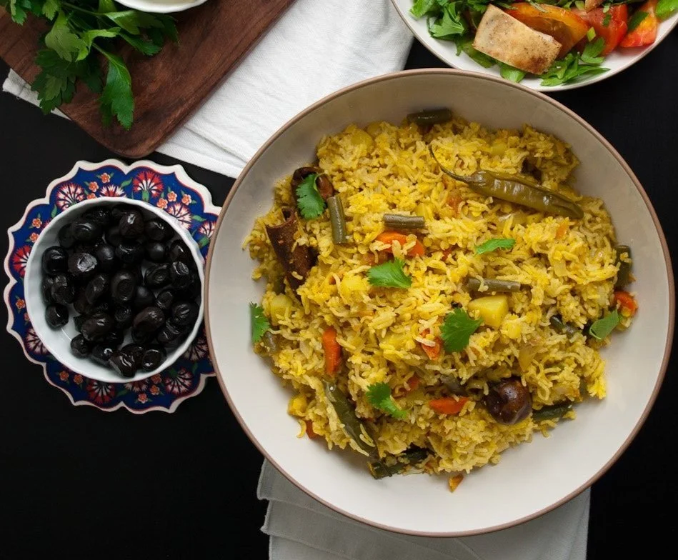 A bowl of yellow rice with vegetables and cilantro, a small bowl of black olives, and a plate of salad with greens and tofu, all arranged on a dark surface.