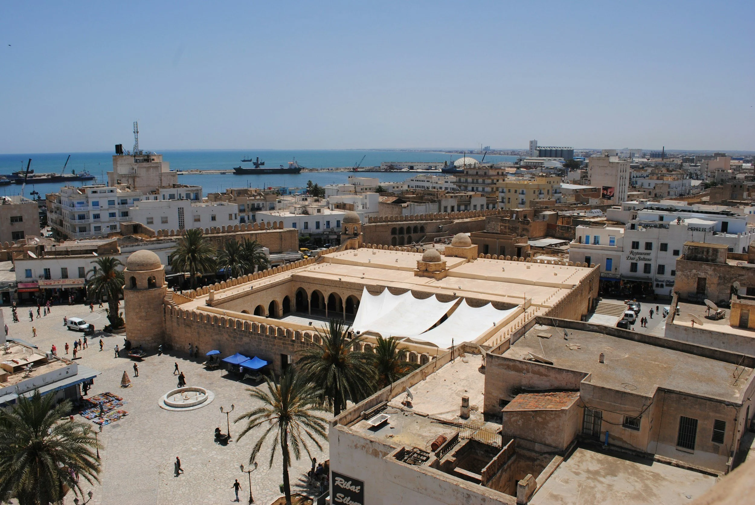 A historic fortress with a tan stone wall and domed towers overlooking a busy city square with palm trees, marketplace stalls, and a fountain, with the ocean in the background and ships docked at the port.