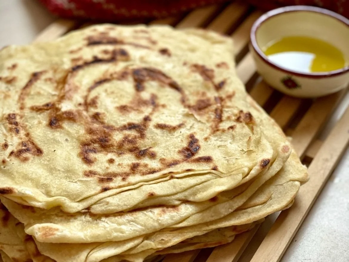 Stack of cooked flatbreads on a wooden board with a small bowl of olive oil in the background.