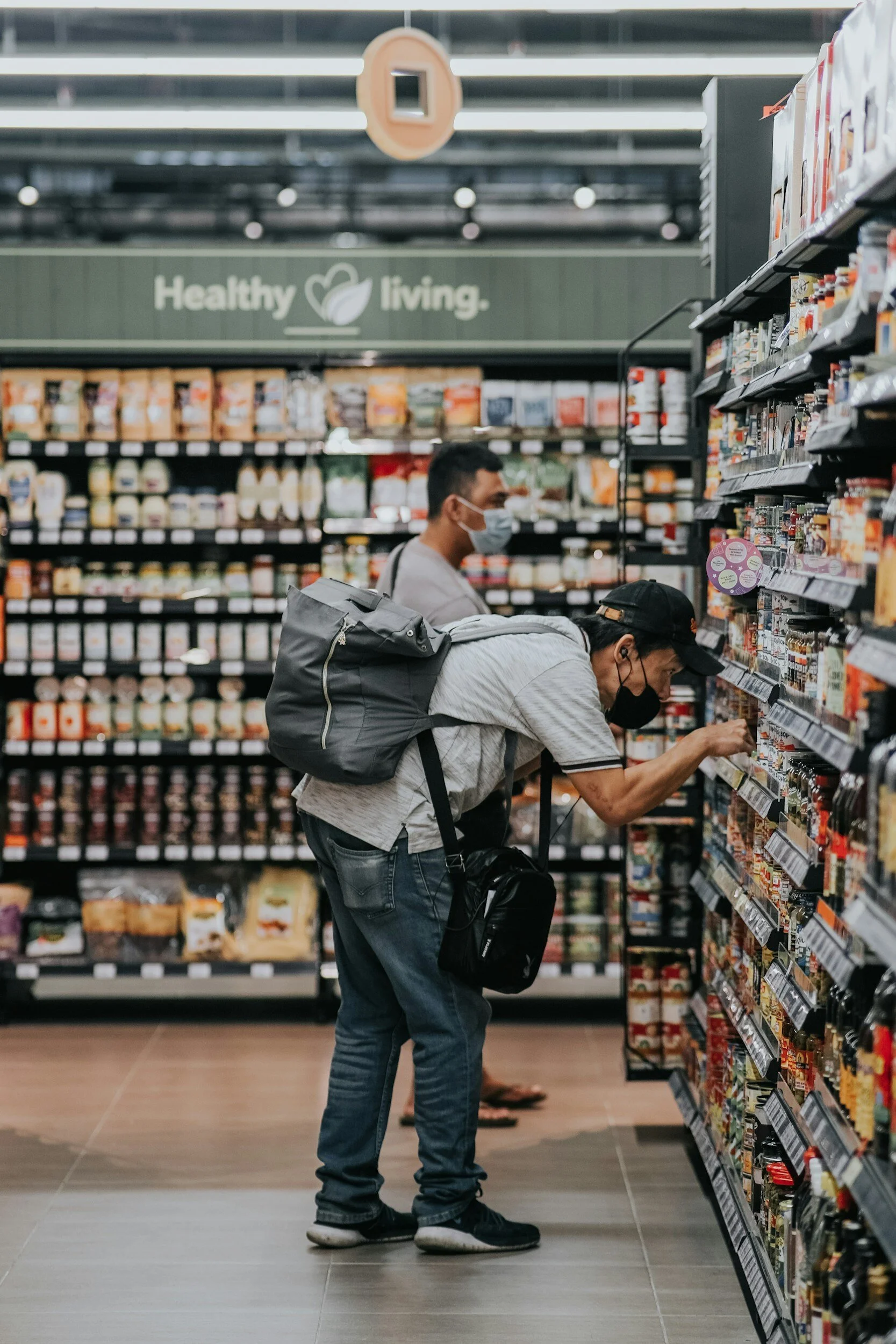 Two men wearing face masks shopping for groceries in a store aisle, one is bent over looking at products on the shelf, and the other stands behind him. The store has a sign that says 'Healthy living'.