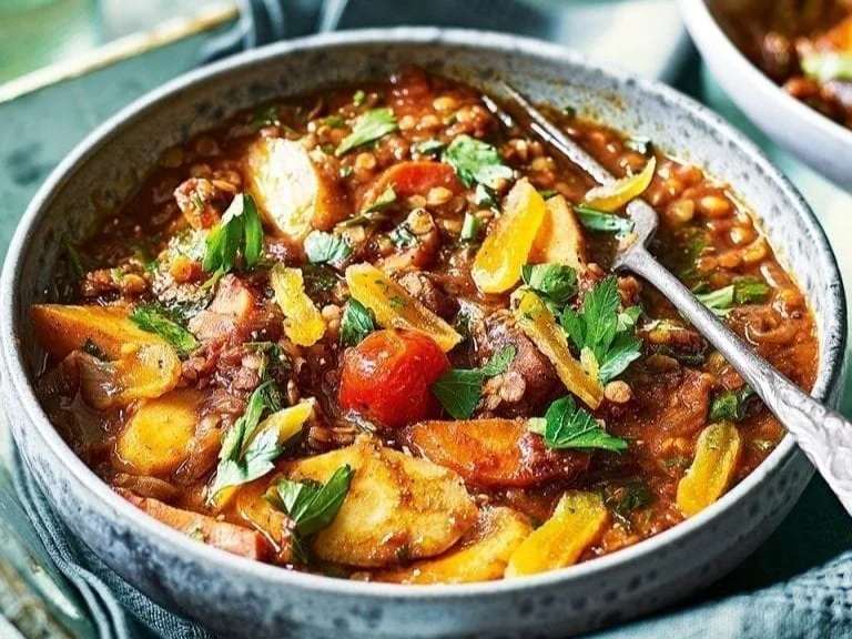 A bowl of vegetable stew with potatoes, carrots, celery, yellow and red bell peppers, garnished with parsley.