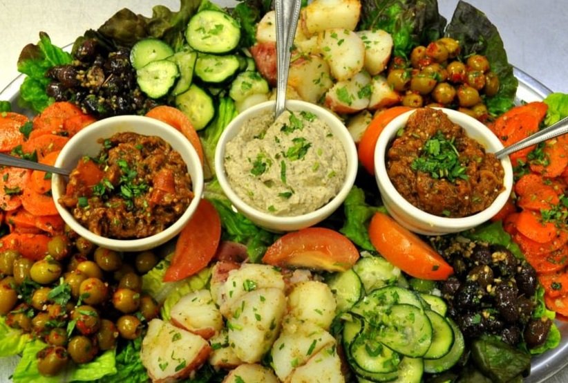 A large platter of assorted Mediterranean-style appetizers including hummus, tabbouleh, roasted vegetables, cucumber slices, and potato salad, garnished with fresh herbs and served with small bowls of different dips.