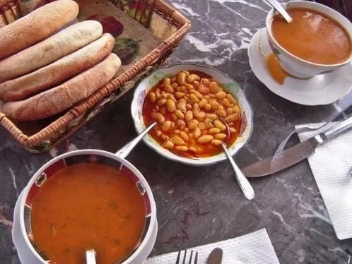 A meal on a marble table with a basket of bread, two bowls of soup, a bowl of beans, a fork, and a spoon.