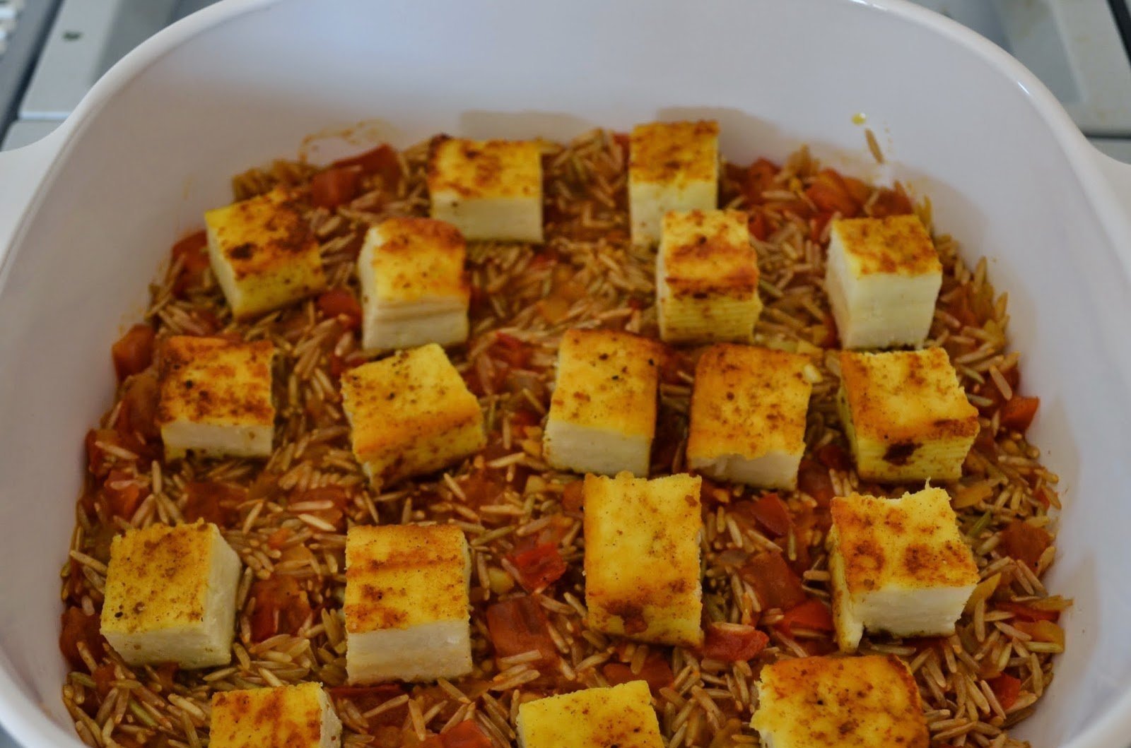 Baked rice dish with diced tomatoes and tofu cubes sprinkled with seasoning in a white baking dish.