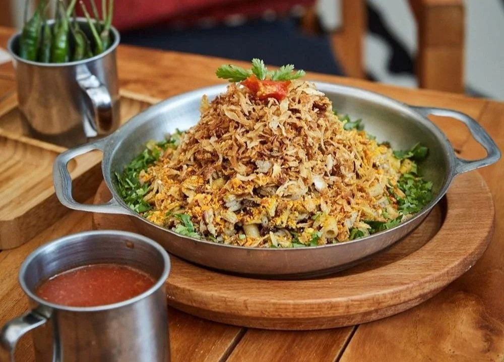 A skillet filled garnished with a sprig of cilantro, served on a wooden platter with side cups of sauce and jalapeños in a small metal cup.