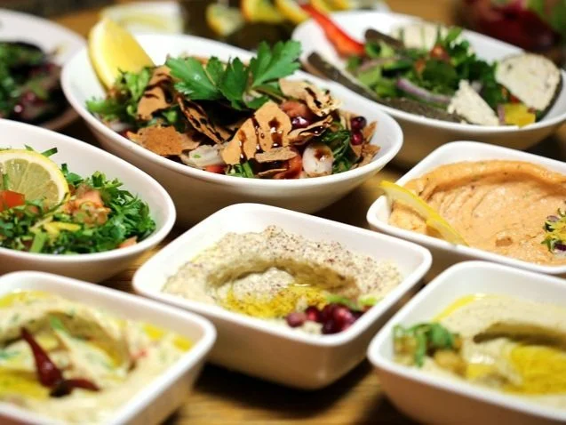 Assorted Middle Eastern dips and salads on white plates, including hummus, baba ganoush, tabbouleh, and falafel with garnishes like lemon and parsley.