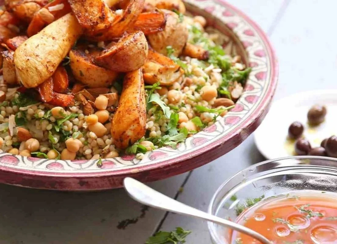 A colorful plate with couscous and roasted vegetables, garnished with herbs. Side dishes include small bowls with olives and a bowl of tomato-based sauce.