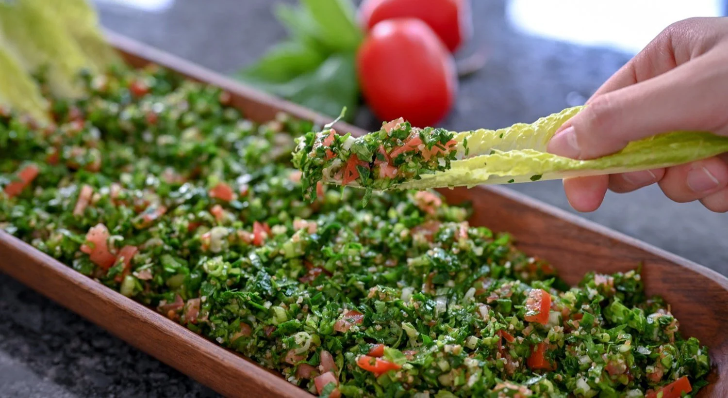 Close-up of a hand holding a lettuce leaf with tabbouleh salad, with a wooden tray of tabbouleh and tomatoes in the background.