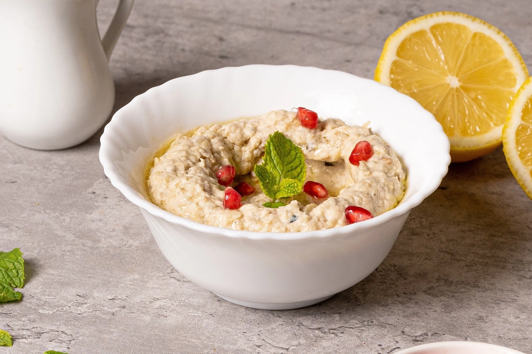 Bowl of hummus garnished with pomegranate seeds and mint leaves, with lemon halves and a white pitcher in the background.
