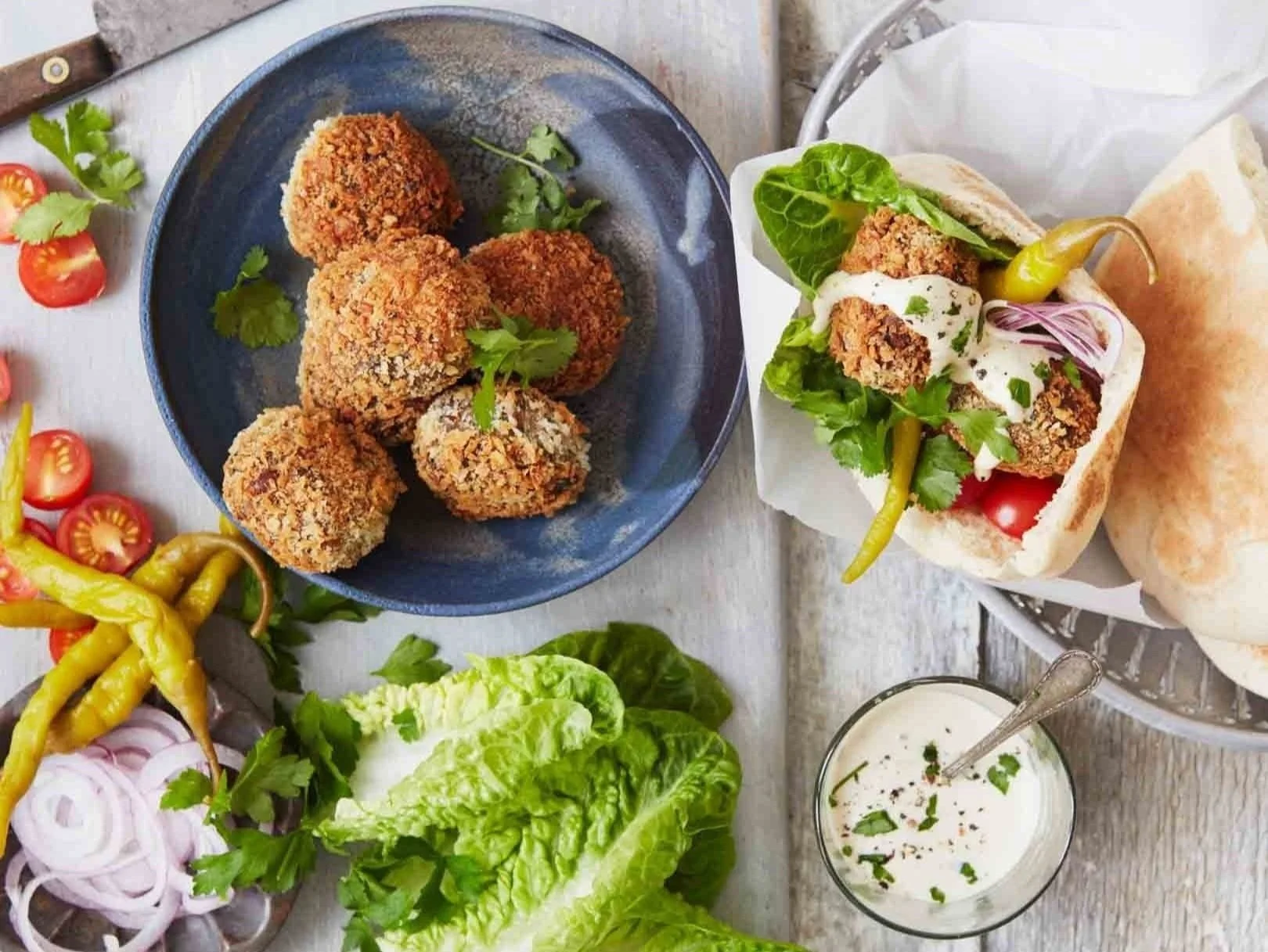 Fried falafel balls on a blue plate, falafel sandwich with greens and dressing, fresh cherry tomatoes, sliced onions, yellow and green peppers, lettuce, and a glass of white sauce with herbs on a white wooden table.