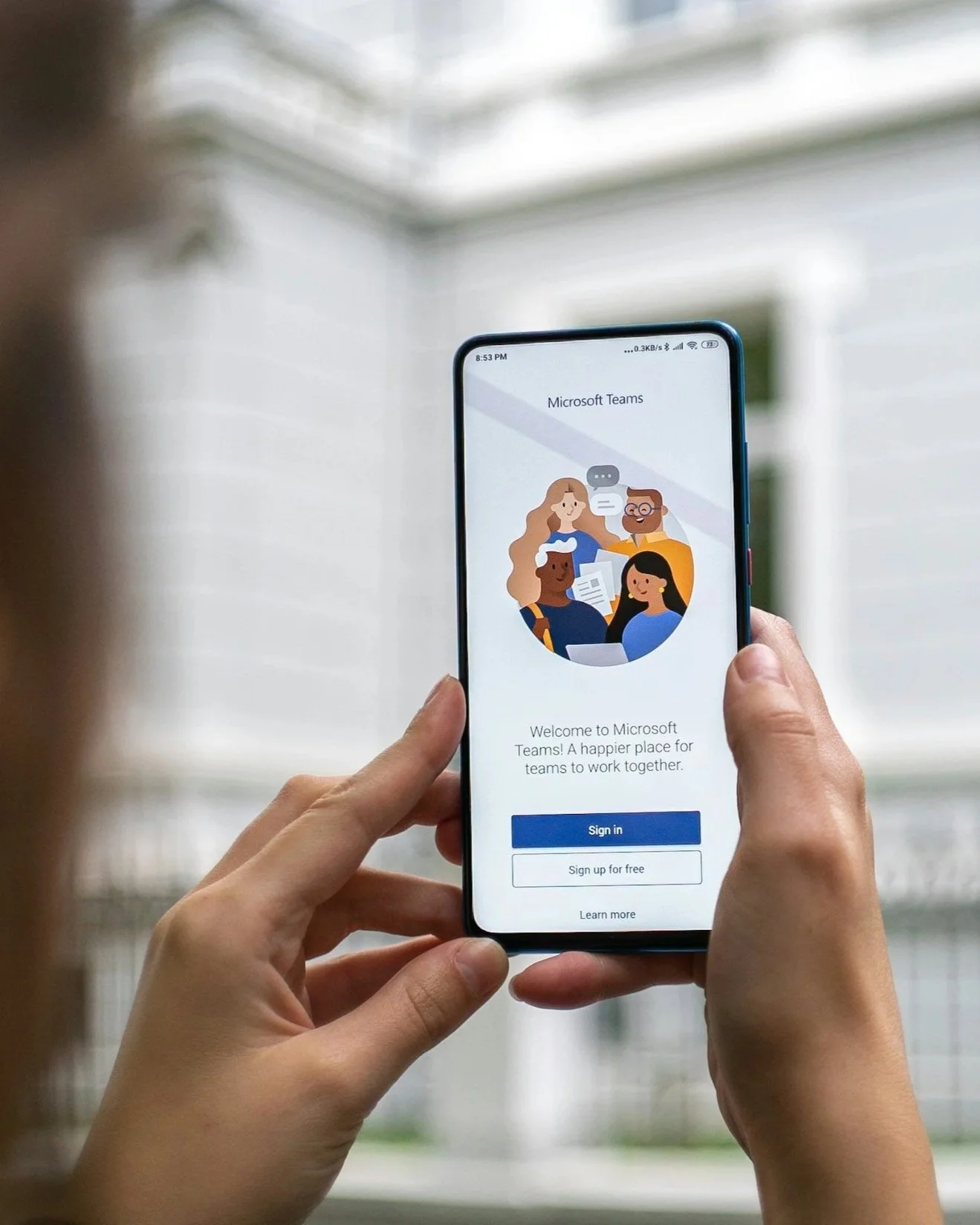 En person håller i en smartphone som visade startskärmen för Microsoft Teams-appen, med knappar för att logga in och registrera sig gratis.