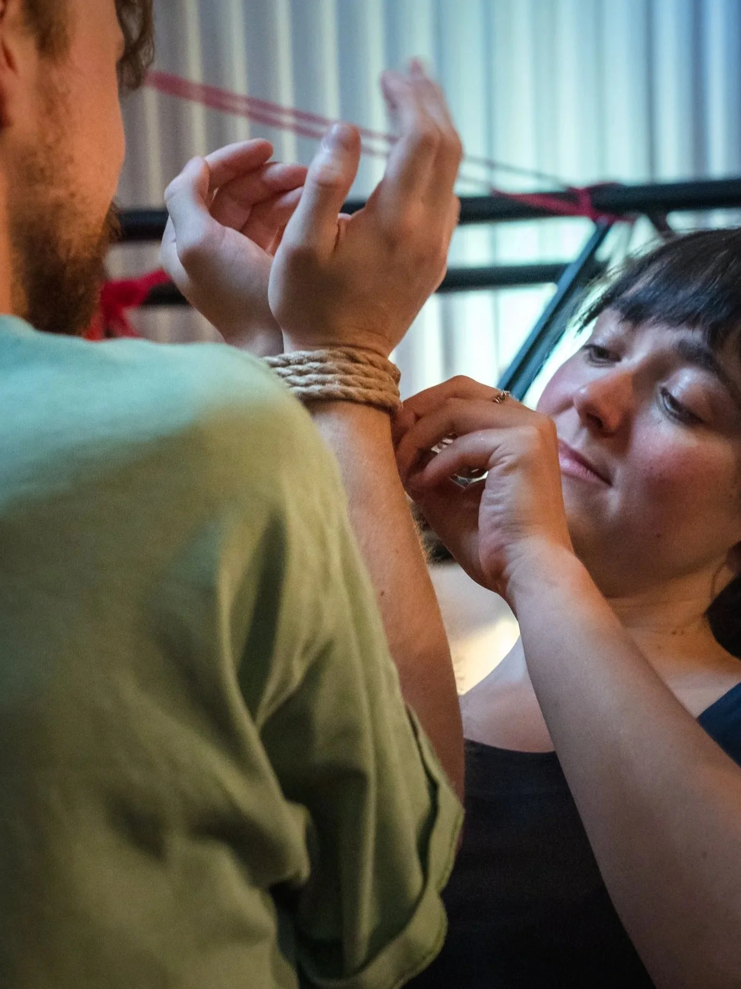 A woman is tying a rope around a man's wrist, who has his arm raised. They are indoors near a curtain, with the woman's focused on tying the knot.