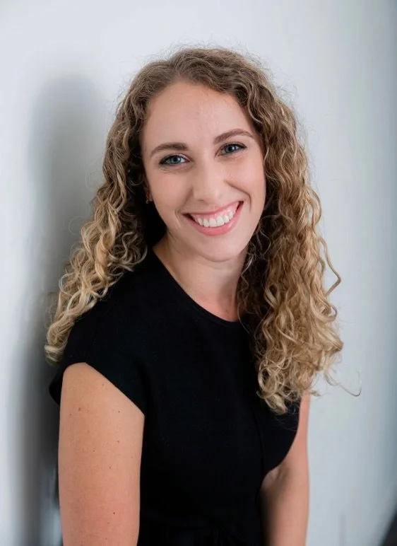 A young woman with curly blonde hair and a bright smile, wearing a black top, standing against a plain light-colored wall.
