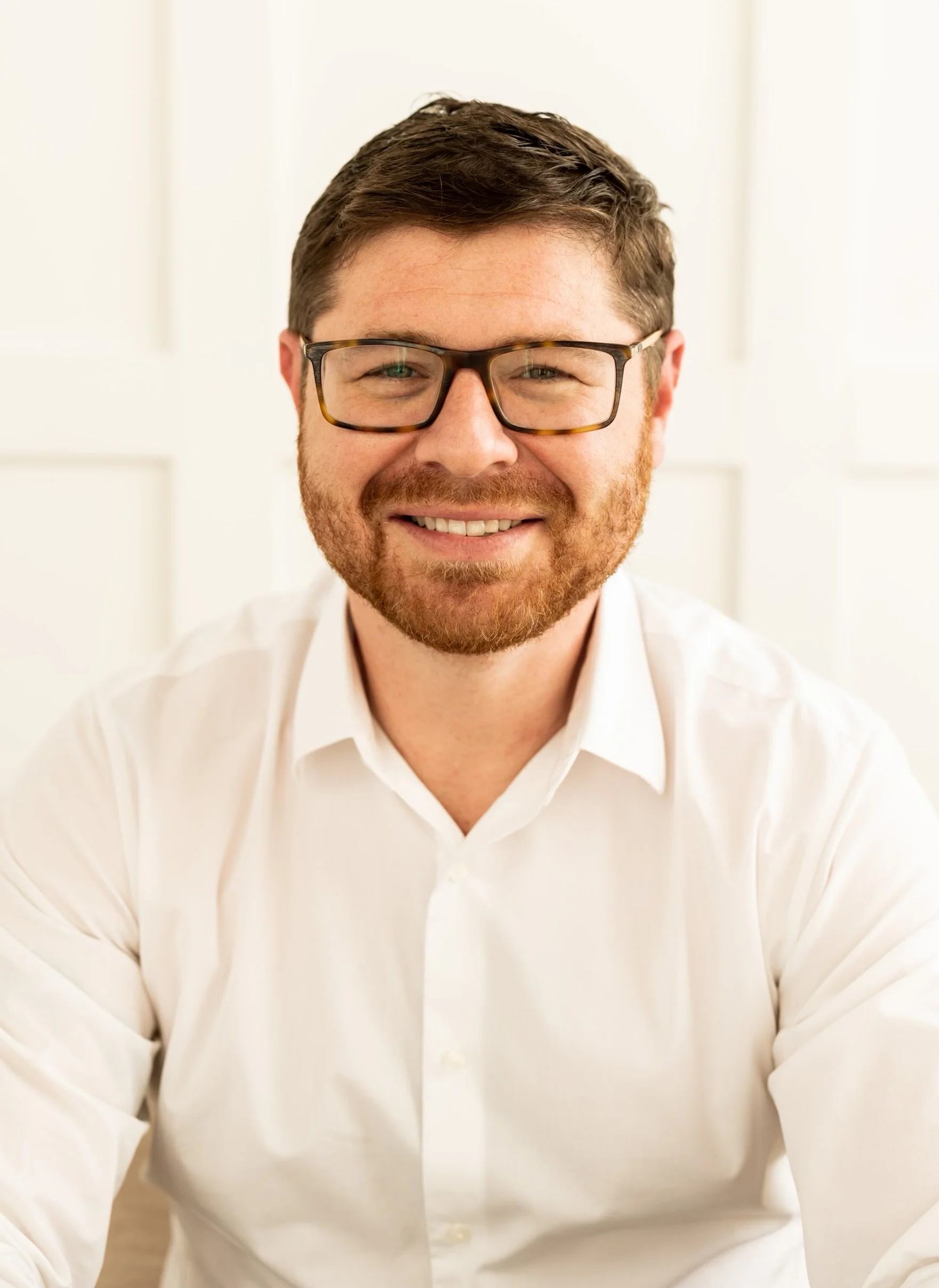 A smiling man with glasses and a red beard wearing a white shirt, set against a white background.