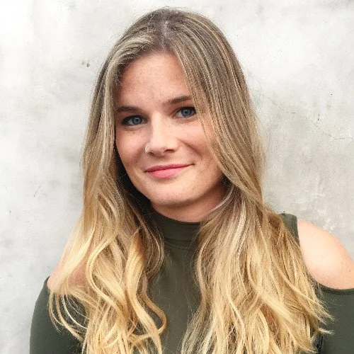 Portrait of a young woman with long blonde hair, blue eyes, wearing a green top with cold shoulder cutouts, standing against a plain light gray background.